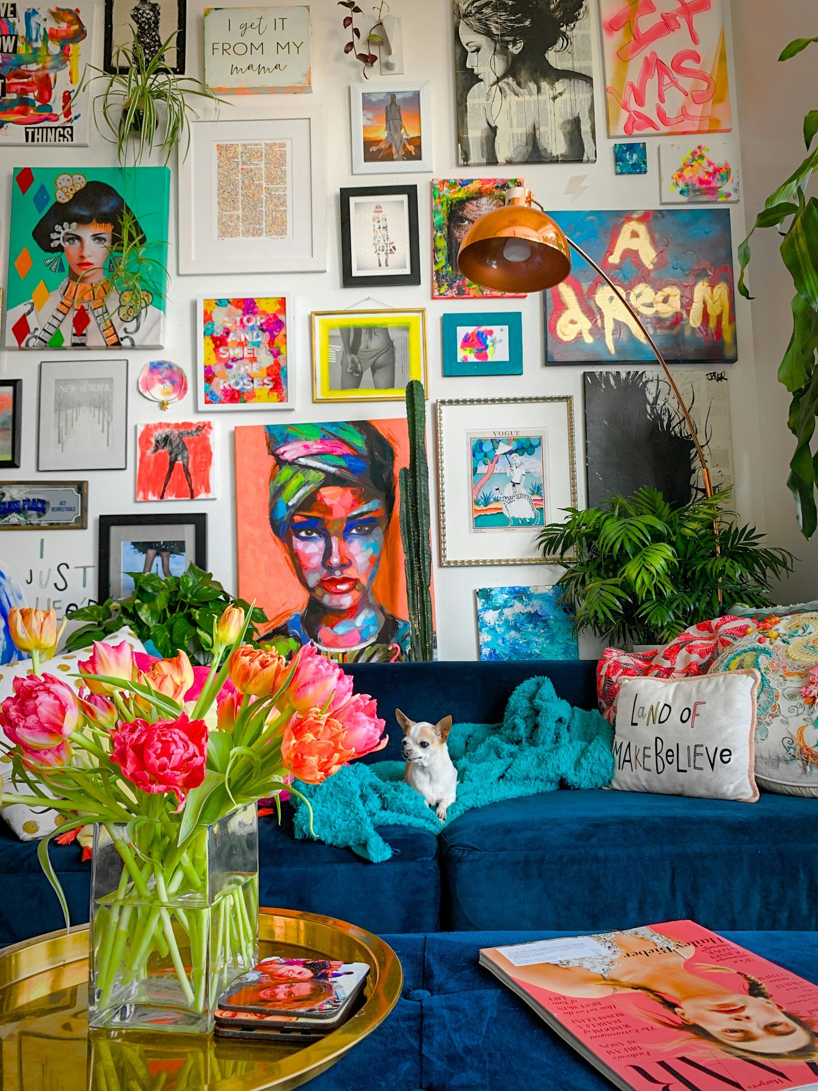 A floor-to-ceiling gallery wall of colourful portraits and prints above a navy velvet sofa with patterned cushions, a small dog on a teal throw, pink tulips and a brass coffee table